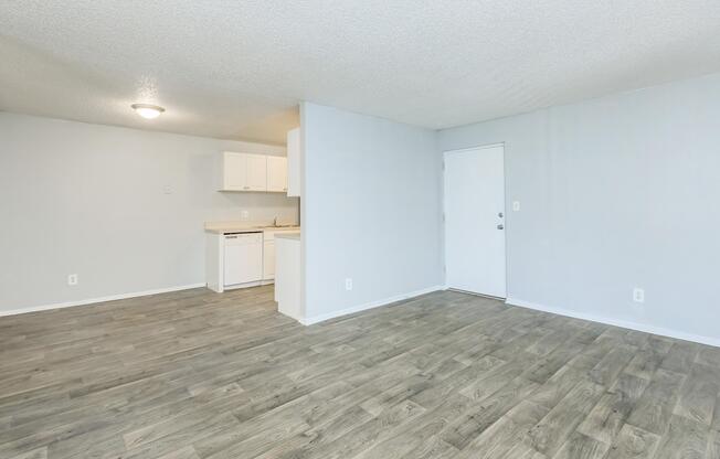 A small, empty living space features a light gray wall and wooden flooring. On the left, there is a kitchenette with white cabinets and a countertop. A door on the right leads outside, while overhead lighting brightens the room. The overall atmosphere is clean and ready for decoration.
