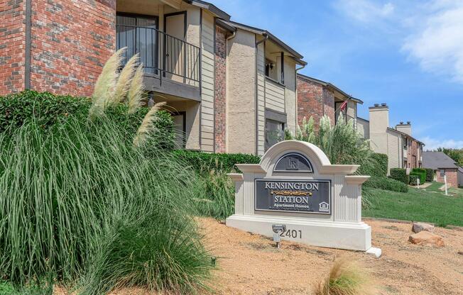 Sign for "Kensington Station" at the entrance of an apartment complex, featuring a landscaped area with tall grass and brick buildings in the background. The sign displays the address "2401" and is surrounded by well-maintained greenery under a clear blue sky.