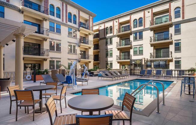 A tranquil pool area featuring lounge chairs and tables, surrounded by modern apartment buildings. Sunlight reflects off the water, with decorative elements like potted plants enhancing the inviting atmosphere. Ideal for relaxation and leisure.