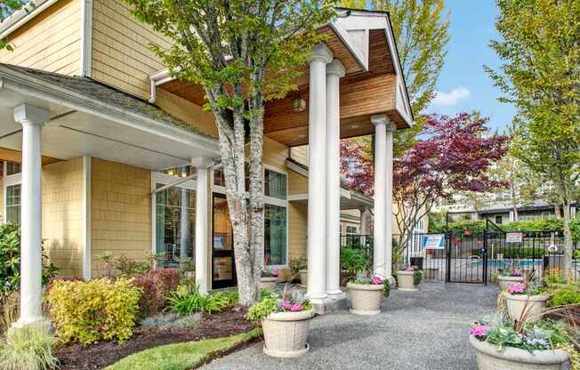 A welcoming community entrance here at Portsmith featuring a covered entryway with tall white columns, light-colored exterior siding, large windows, and landscaped planters filled with seasonal flowers, with mature trees and a gated amenity area visible along a paved walkway that creates an inviting and well-kept first impression.
