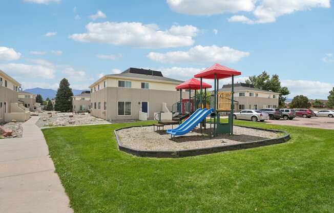 A playground with a blue slide and red canopy is surrounded by a grassy area and apartment buildings.