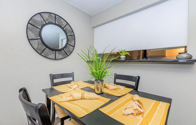 A modern dining area featuring a black table set with yellow placemats and folded napkins. A small green plant in a decorative pot sits in the center. A round mirror with a geometric design hangs on the wall, and there is a partial view of a kitchen area in the background.