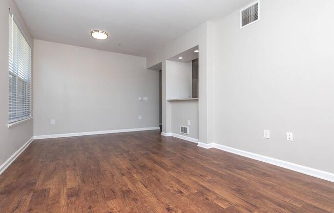 A spacious, empty living room with light-colored walls and a wooden floor. A large window allows natural light, and there's a ceiling light fixture. To one side, a partial view of a kitchen area can be seen, featuring a bar space, but the overall ambiance remains open and uncluttered.