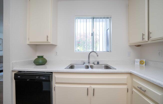 Modern kitchen with light-colored cabinets, a double sink, and a small green decorative vase. The countertop is made of speckled material, and there's a window above the sink letting in natural light. A black dishwasher is visible on the left side of the sink area.