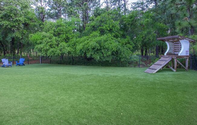 A spacious green backyard featuring artificial turf, surrounded by trees. On one side, there are two blue lounge chairs, and on the other, a wooden climbing structure for pets with a ramp leading up to a small playhouse. The scene is tranquil and well-maintained, ideal for outdoor activities.