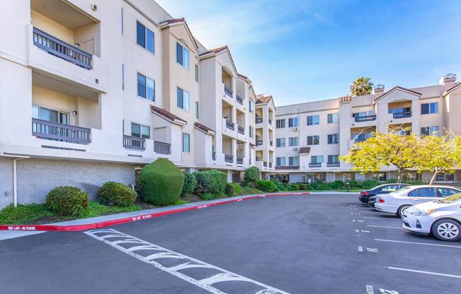 A residential apartment building with a cream-colored exterior and balconies, surrounded by well-maintained landscaping. In the foreground, there is a painted parking lot with several parked vehicles and a tree, against a clear blue sky. The setting reflects a suburban environment.