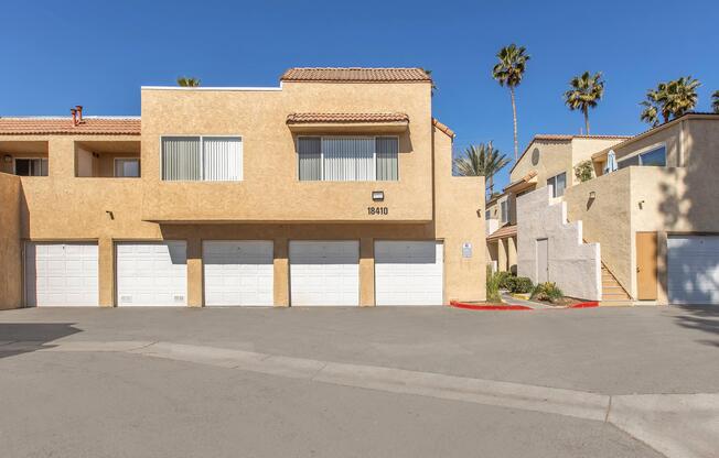 A well-maintained residential building with a light brown stucco exterior. The structure features multiple garage doors and large windows with blinds. The scene is sunny, with a clear blue sky and several palm trees in the background, creating a warm, inviting atmosphere. The surrounding area is paved.