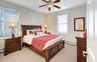A cozy staged bedroom here at Prairie Pines at the Ridge featuring a dark wood bed with built-in storage drawers, neutral carpeting, matching nightstands with lamps, large windows that bring in natural light, and a ceiling fan that adds comfort and airflow.