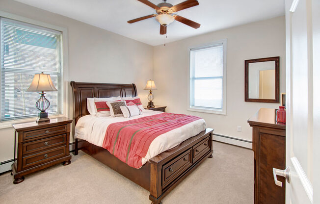A cozy staged bedroom here at Prairie Pines at the Ridge featuring a dark wood bed with built-in storage drawers, neutral carpeting, matching nightstands with lamps, large windows that bring in natural light, and a ceiling fan that adds comfort and airflow.