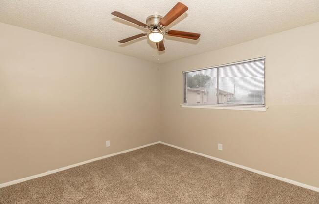A neutral-toned empty room featuring a ceiling fan with wooden blades, a window with blinds allowing natural light, and beige carpet flooring. The walls are painted in a soft cream shade, giving the space a bright and inviting feel. Ideal for customization or decoration.