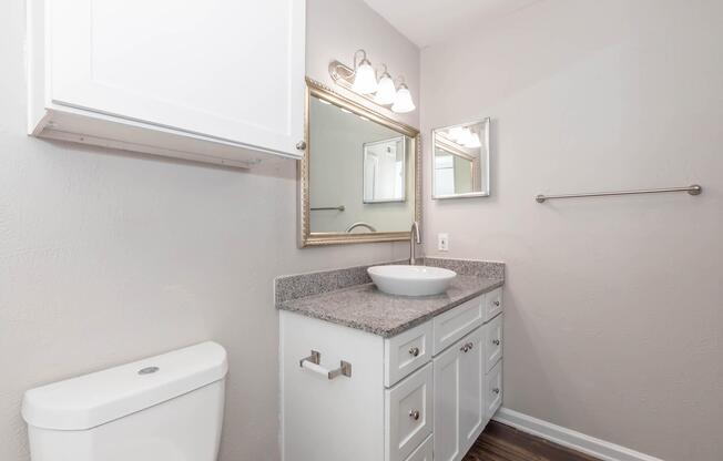 A modern bathroom featuring a white vanity with a granite countertop, a round sink, and a large mirror framed in gold. The walls are painted light gray, and there is a toilet in the corner. Soft lighting is provided by wall-mounted fixtures above the mirror. The flooring is dark wood.