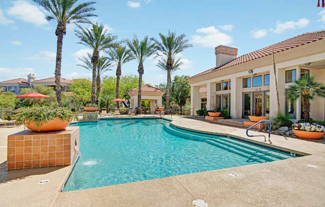 A resort-style swimming pool here at Mirador surrounded by tall palm trees, terracotta tile accents, and spacious sun decks. The sparkling blue water curves alongside wide entry steps and stainless handrails, while vibrant planters, shaded seating areas, and a stucco clubhouse with red tile roofing create a warm, inviting atmosphere perfect for relaxing outdoors.