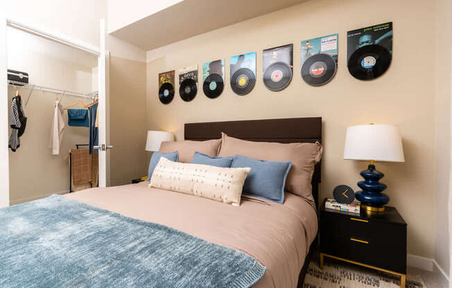 A bedroom with a bed, pillows, a lamp, and a wall with framed records on it.