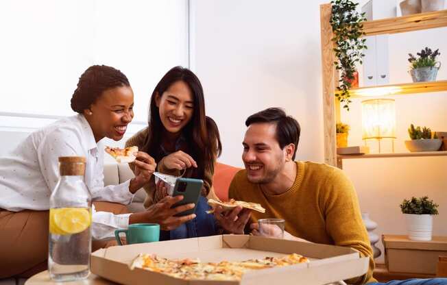 a group of people sitting around a table eating pizza and looking at a cell phone