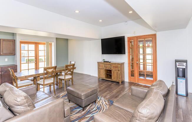 A spacious living area featuring a comfortable seating arrangement with brown leather sofas, a coffee table, and a wooden dining table with chairs. Thereâs a large flat-screen TV mounted on the wall and glass doors leading to an outdoor patio. Natural light fills the room, enhancing the modern decor.