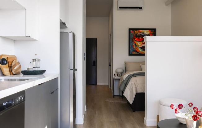 A modern kitchen with a refrigerator, a dishwasher, and a bedroom with a bed and a painting on the wall.