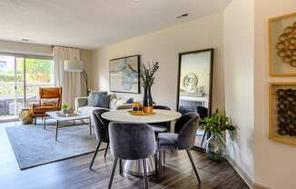 A welcoming open living and dining area here at Retreat at McAlpine Creek featuring wood-style flooring, a round dining table with upholstered chairs, and a comfortable living room setup with a sofa, accent chair, and coffee table. Decorative wall art, greenery, and a floor lamp add style, while sliding glass doors bring in natural light and balcony views.