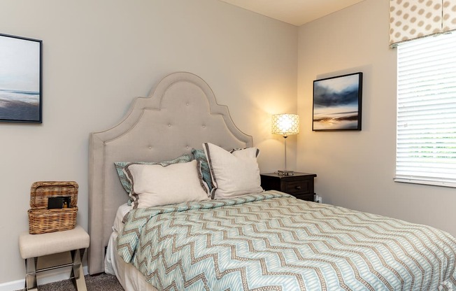 A bed with a blue and white patterned comforter sits in a room with a white wall and a window with blinds.