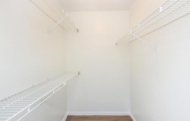 Empty closet with white wire shelving along the walls. The walls and ceiling are painted white, and the floor is wooden. The space is well-lit and uncluttered, providing a blank canvas for organization or storage.