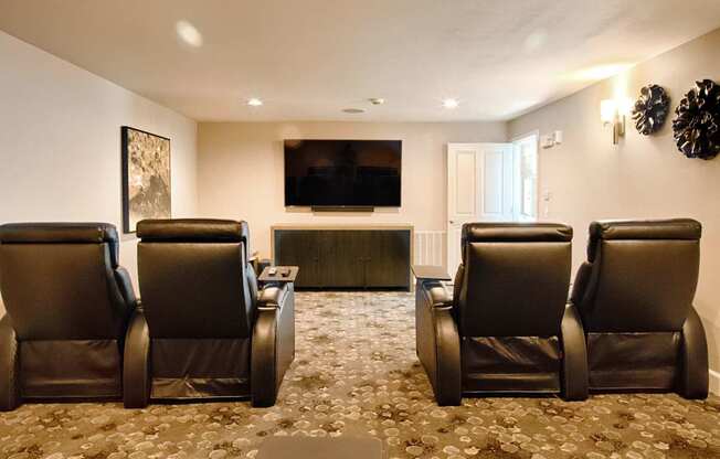 A private theater room here at Andorra featuring plush black reclining chairs arranged in rows, a wall-mounted flat-screen television above a sleek media console, soft patterned carpeting, warm wall sconces, and recessed ceiling lighting that creates a comfortable and immersive movie-watching environment.