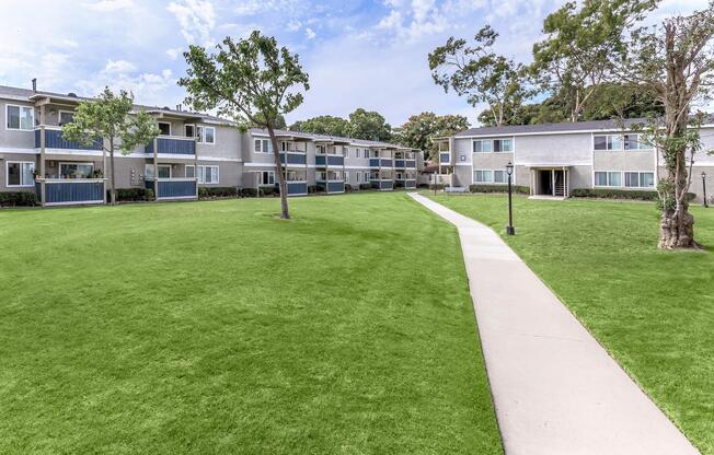 A well-maintained apartment complex with two-story buildings surrounding a manicured lawn. A smooth concrete pathway runs through the green grass, leading to entrances of the apartments. Trees are strategically placed throughout the area, and the sky is partly cloudy, adding to the pleasant atmosphere.
