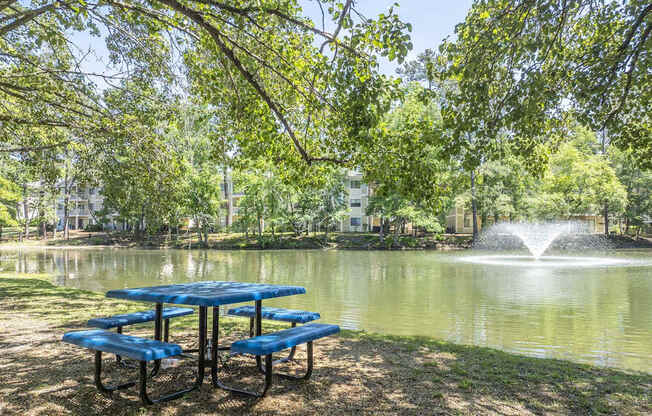 A blue picnic table is set up by a pond with a fountain in the background.