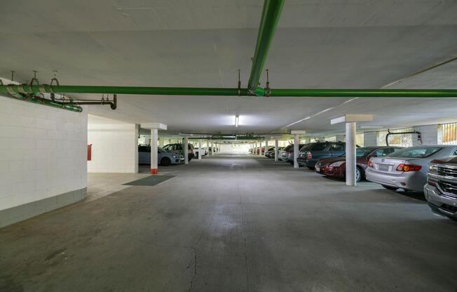 A spacious, well-lit underground parking garage featuring several parked cars on both sides. The concrete floor is visible, along with green pipes running along the ceiling and rows of white columns supporting the structure. The far end of the garage is dimly lit, leading to an exit.