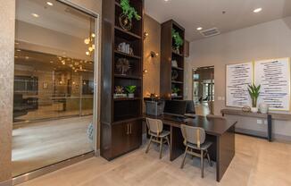 A modern office space featuring a sleek desk setup with two chairs, decorative shelves filled with plants and decorative items, large windows allowing natural light, and a welcoming atmosphere. The design incorporates neutral tones and contemporary decor elements.