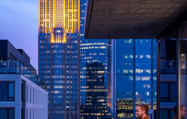 Two people are standing on a balcony with a cityscape at dusk.