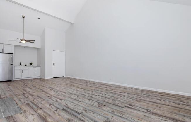 Spacious, minimalist living area with light-colored wooden flooring and high ceilings. A kitchen area with a stainless steel refrigerator is visible in the background. The walls are painted white, creating a bright, airy feel. A single door leads outside, enhancing the open layout.