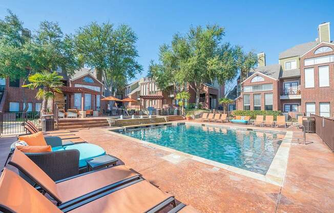 A resort-style outdoor pool here at Lakewood on the Trail with clear blue water, spacious sun deck, lounge chairs, shaded seating areas, and surrounding brick buildings, creating a bright, relaxing community space.