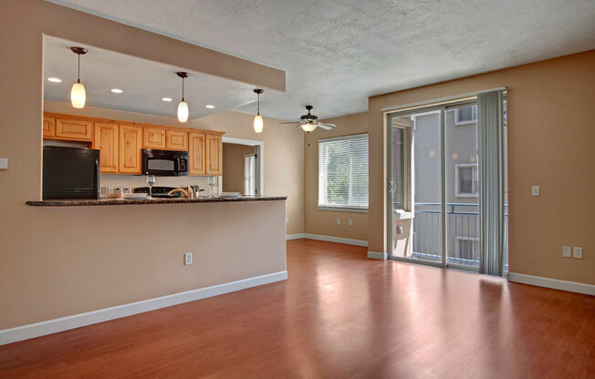 A living and kitchen space here at Bridges with warm wood flooring, light wood cabinets, granite breakfast bar, black appliances, pendant lights, and a bright sliding glass door leading to a private balcony.