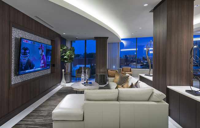 Lounge with dark wood-paneled walls, a large, white leather sectional couch, a flat-screen TV, modern accessories and art, and floor-to-ceiling windows with a city view.