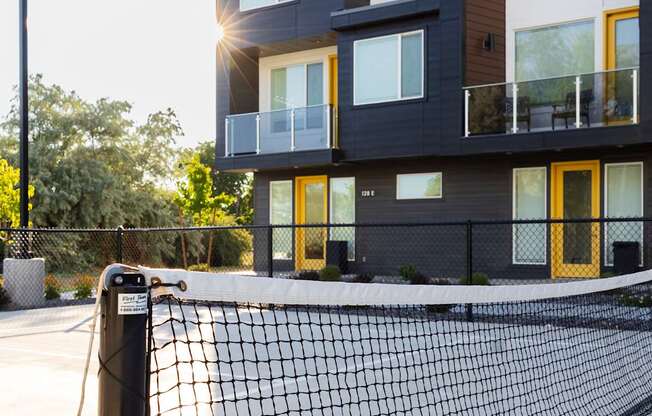 A tennis court with a black net in front of a modern building with balconies.