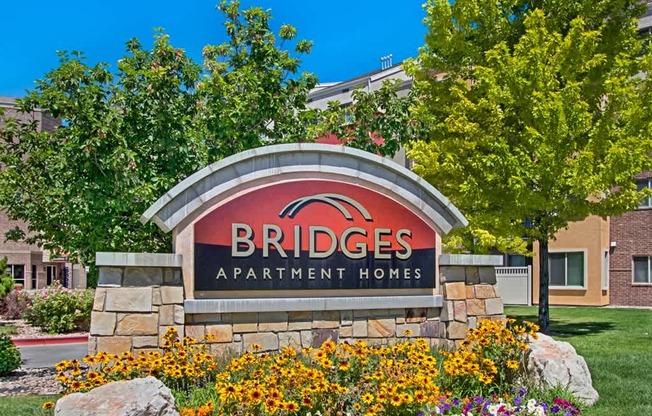 A community monument sign here at Bridges with a stone base and curved red-and-black Bridges Apartment Homes panel, surrounded by bright yellow flowers, green trees, and landscaped greenery under a clear blue sky.