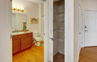 A bright bathroom and entry area here at Prairie Pines at the Ridge featuring a wood vanity with laminate countertop, large wall mirror with overhead lighting, a tub and shower combination, vinyl plank flooring, a nearby linen closet with shelving, and a welcoming entry door that adds everyday functionality and storage.