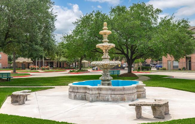 A decorative stone fountain sits at the center of a grassy area, surrounded by trees. The fountain features multiple tiers and is filled with blue water. Two stone benches are placed nearby on a concrete surface, with a peaceful park setting in the background. Bright blue sky and fluffy clouds add to the serene atmosphere.