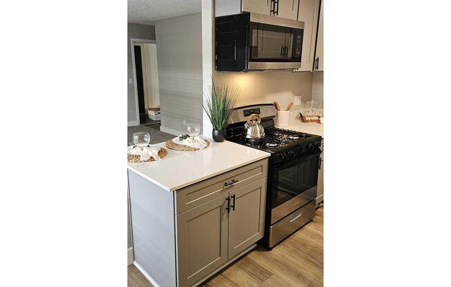 A modern kitchen featuring light-colored cabinetry, a stainless steel gas stove, and a microwave above. The countertop is white with a decorative tray, glass dish, and plant. The flooring is wooden and there is a glimpse of adjacent living space in the background.