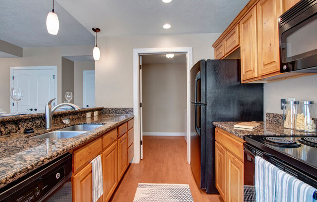 A kitchen here at Bridges with granite countertops, warm wood cabinets, black appliances, pendant lights, a double sink, and a long breakfast bar overlooking the open living space.