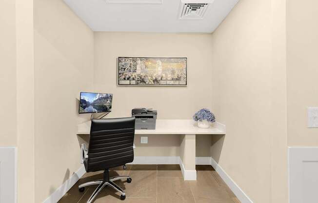 A black office chair is in front of a desk with a computer monitor and a framed picture on the wall.