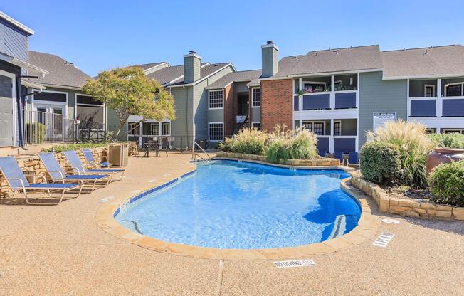 A sunlit swimming pool surrounded by lounge chairs and landscaped greenery, with apartment buildings in the background. The pool has a curved shape and is set in a residential area, creating a relaxing atmosphere for residents.
