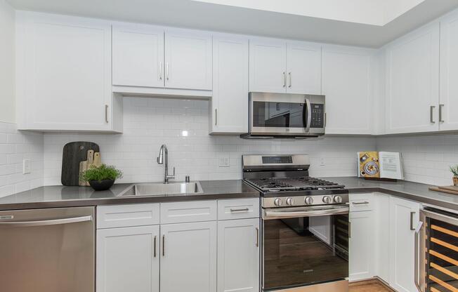 A modern kitchen featuring white cabinetry, a stainless steel gas stove, a microwave, and a sleek countertop. There is a stainless steel sink with a single handle faucet, a wooden cutting board on the counter, and a small potted plant. The flooring is wooden, adding warmth to the contemporary design.