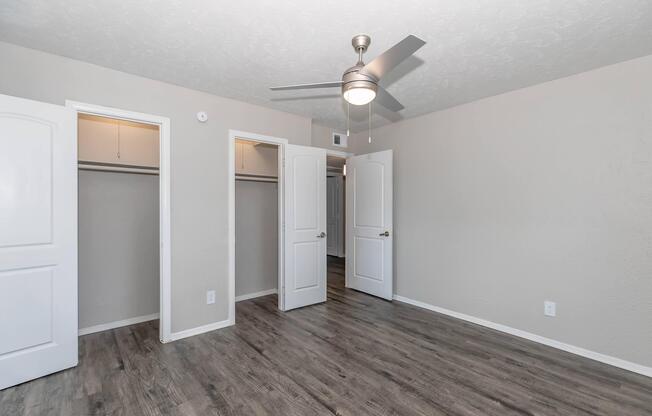 A spacious bedroom featuring light-colored walls and wood-look vinyl flooring. The room has two open doors leading to closets, a ceiling fan, and a neutral color palette, creating a bright and inviting atmosphere.