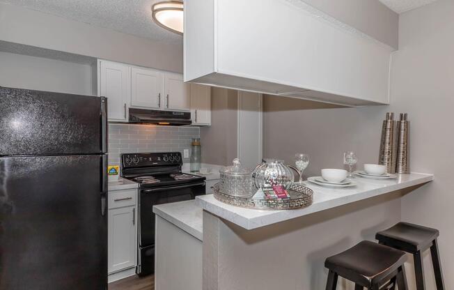 A modern kitchen featuring a black refrigerator, stovetop, and white cabinets. A bar counter with two black stools is in the foreground, showcasing decorative serving dishes and glasses. The walls are painted in neutral tones, and the space is well-lit with overhead fixtures.