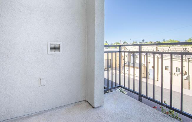 A view from a balcony, featuring a concrete wall with a vent and a railing. Below, there is a glimpse of a courtyard or parking area with several buildings. The sky is clear and blue. Flowers are visible in pots along the edge of the balcony.