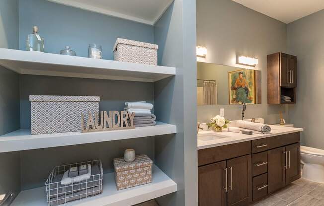 The Residences at Crosspoint laundry nook shows built-in white shelves holding decorative boxes, glass jars, and folded towels alongside a wooden "Laundry" sign, with a bathroom vanity visible in the background.