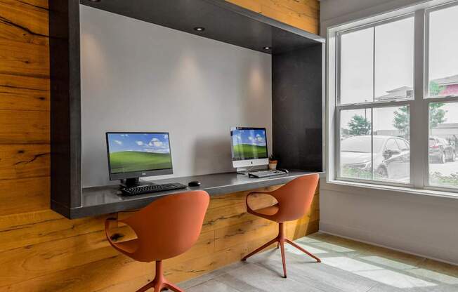 Two computers are on a desk with chairs.