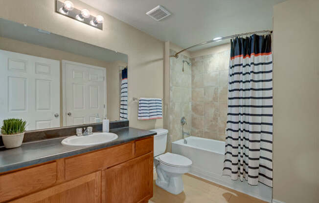 A bright bathroom here at Mission Green showcasing a wide vanity with a wood-tone cabinet, a large wall mirror with overhead lighting, a clean tub and shower combo with tile surround, light neutral walls, and coordinated fixtures that create a fresh, functional, and well-lit space designed for everyday comfort and convenience.