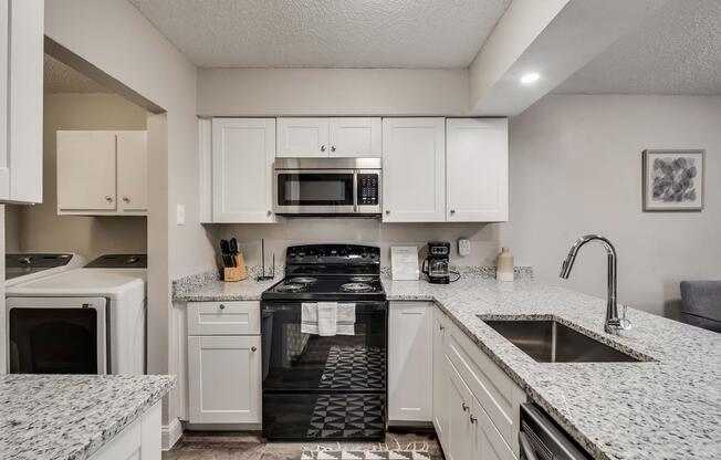 Modern kitchen featuring white cabinetry, granite countertops, a stainless steel oven and microwave, and a black stove. A double sink is positioned in the island, which includes a dish rack in front. Thereâs a laundry area visible to the left, and a coffee maker on the counter. Decor includes a framed wall art piece.