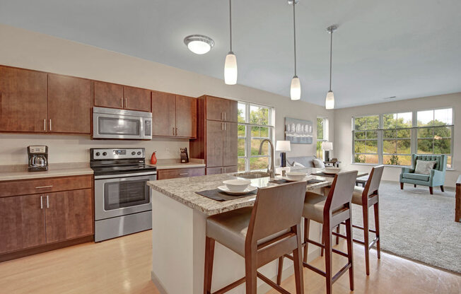 A modern open kitchen and living area here at Vue at Pinnacle Park with warm wood cabinets, stainless steel appliances, granite island with bar seating, pendant lighting, and large windows bringing in bright natural light.